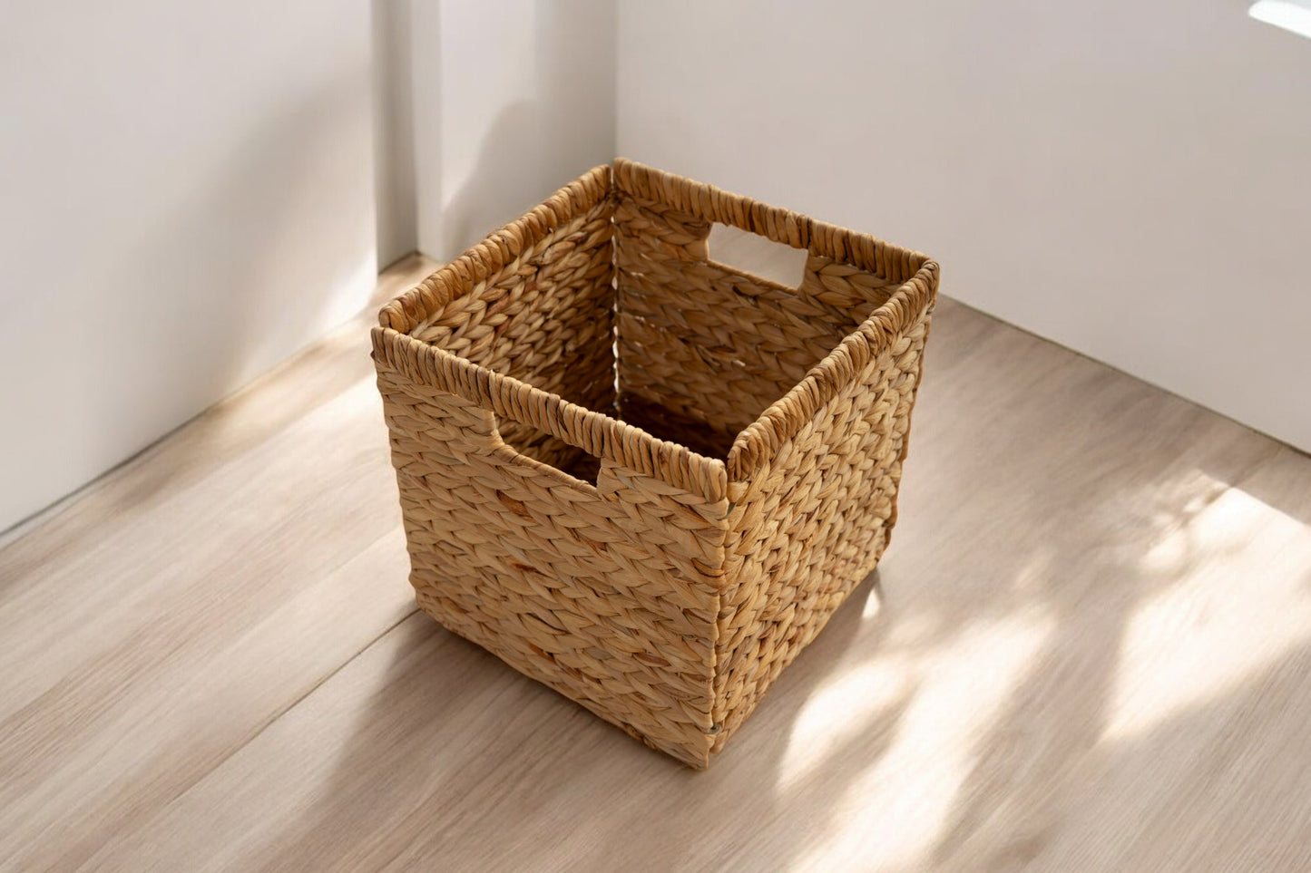 A square woven basket with handles placed on a sunlit wooden floor, perfect for storage and organization.