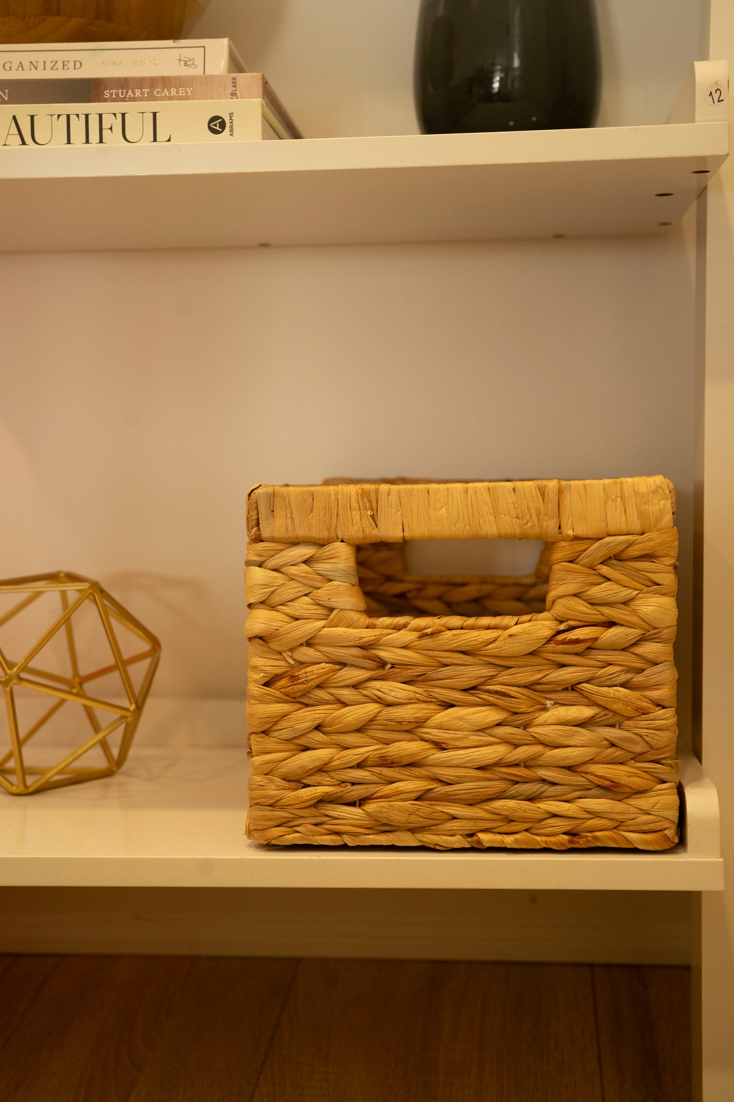 Woven wicker basket with handles placed on a white shelf, surrounded by books and decorative items.