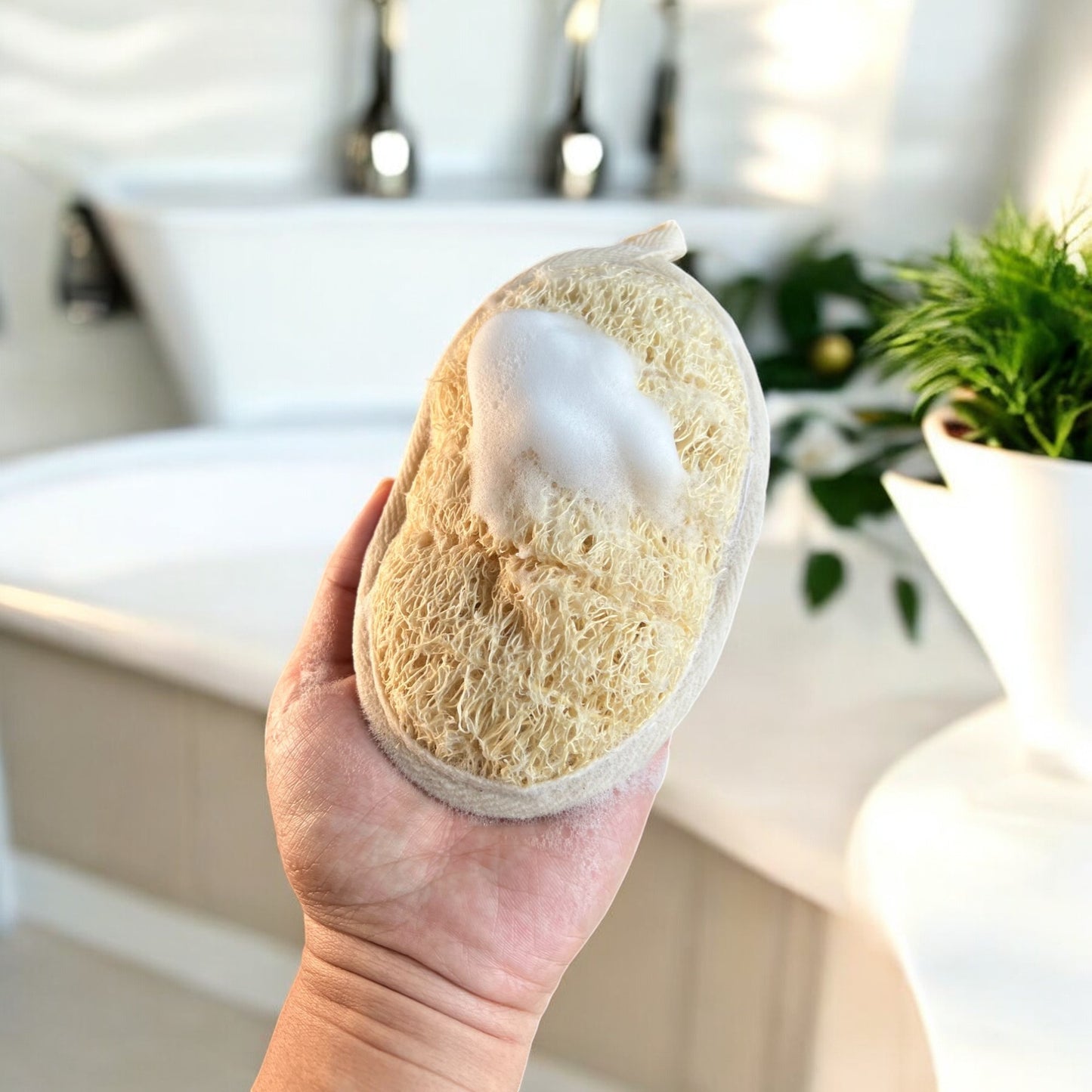 A hand holding an oval-shaped natural loofah sponge with foam on its surface, set in a bathroom environment with a white bathtub and green plants in the background, emphasizing its use for gentle cleansing and exfoliation.