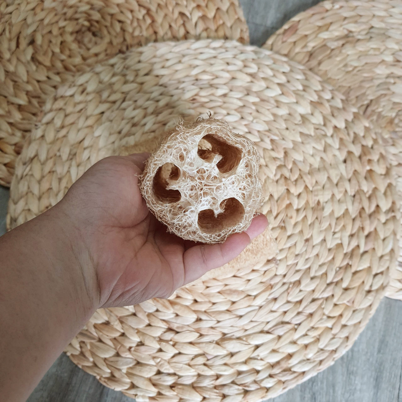 A hand holding a natural loofah sponge, showcasing its fibrous and porous texture, with woven straw mats in the background. Ideal for exfoliation, skincare, and eco-friendly usage
