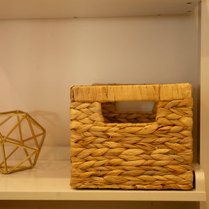 Woven wicker basket with handles placed on a white shelf, surrounded by books and decorative items.